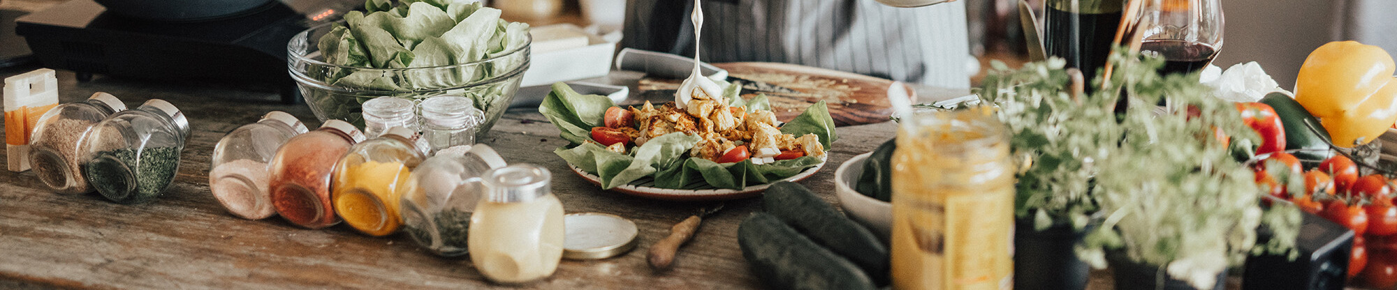 ein Salat, welcher mit einem Dressing übergossen wird. Links befinden sich in Glasbehältern mehrere Gewürze