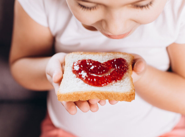 A child is holding a slice of bread with jelly spread in the shape of a heart.